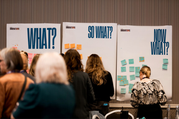 Photo of a group of peope standing around a whiteboard with lots of post-it notes on it at Hauora hinengaro conference.